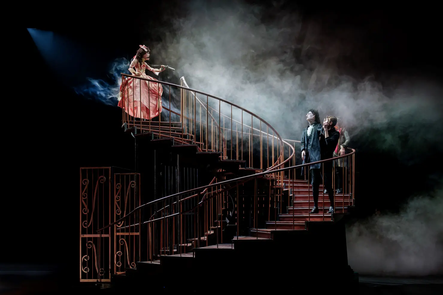 Theateraufführung mit einer dramatischen Treppe. Mehrere Personen stehen oben, während ein Puppenspieler und eine Puppe unten stehen. Nebel und Lichtakzente verstärken die Atmosphäre.