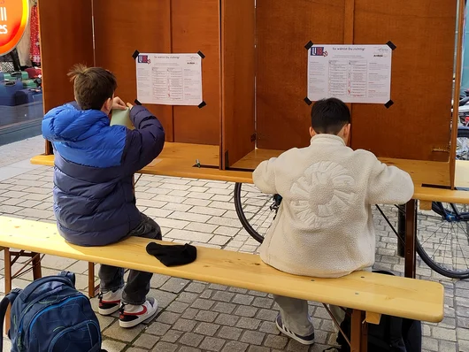 Zwei junge Wähler sind von hinten an der Wahlurne zu sehen. Klick öffnet die Meldung.