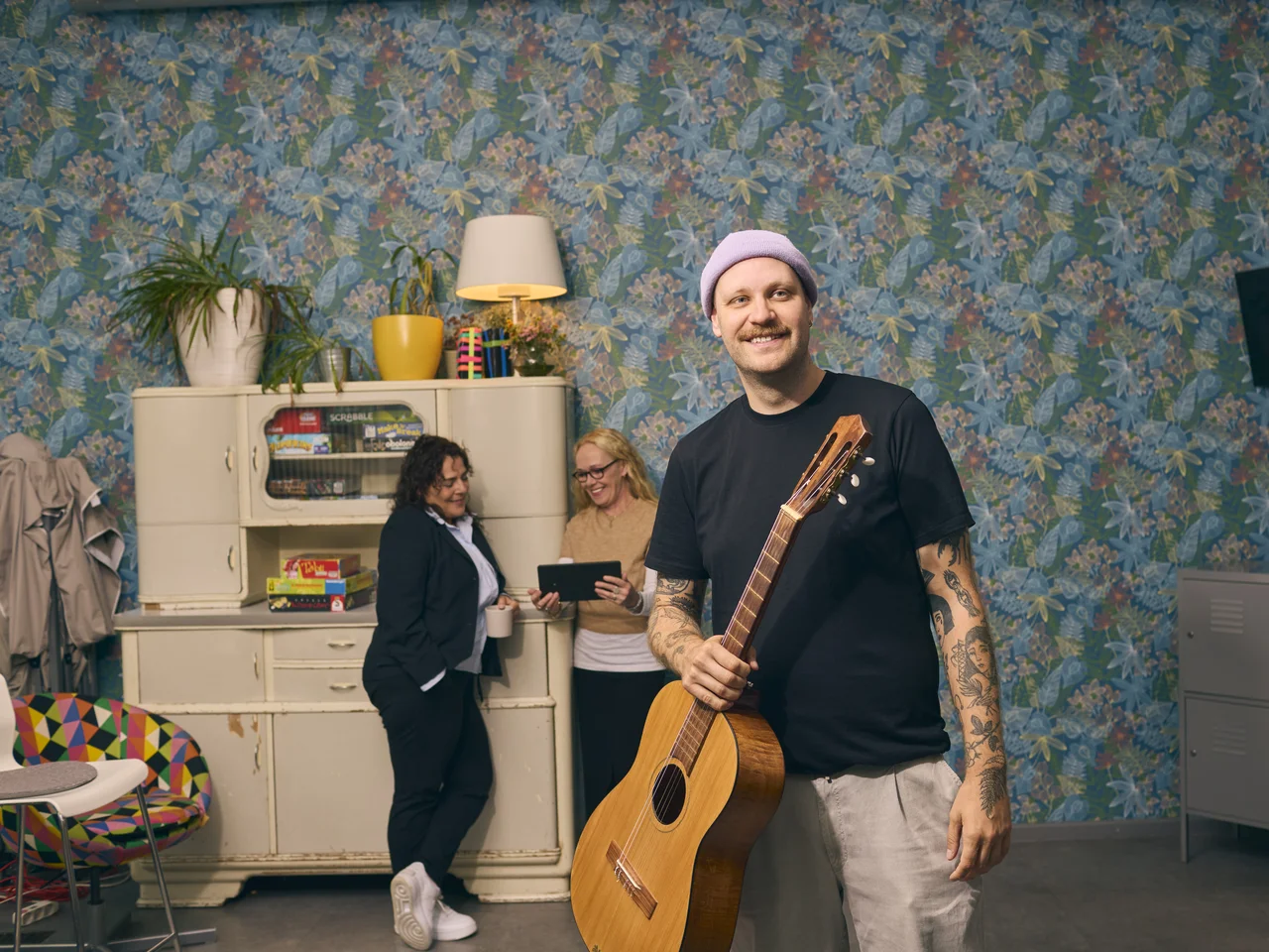 Mann mit Tätowierungen und Gitarre steht vor zwei Frauen, die an einem Schrank mit Spielen ein Tablet betrachten.