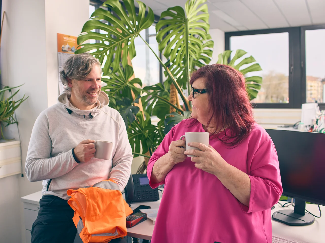 Ein Mann und eine Frau sitzen in einem hellen Büro mit großen Fenstern und grünen Pflanzen und halten jeweils eine Tasse in der Hand und schauen sich an. 