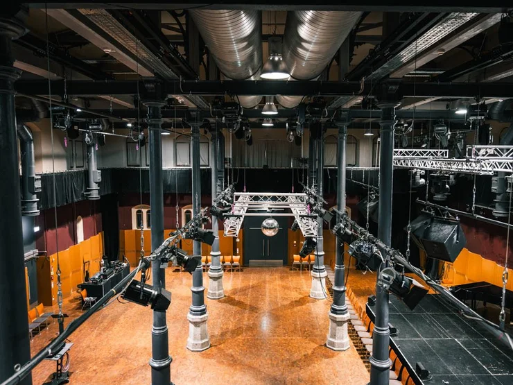 Große Halle des Kulturforums mit Holzboden, schwarzen Säulen und einer zentralen Beleuchtungs- und Lautsprecheranlage an der Decke.