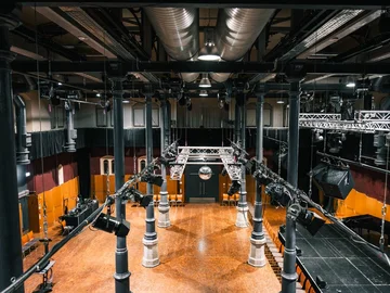 Große Halle des Kulturforums mit Holzboden, schwarzen Säulen und einer zentralen Beleuchtungs- und Lautsprecheranlage an der Decke.