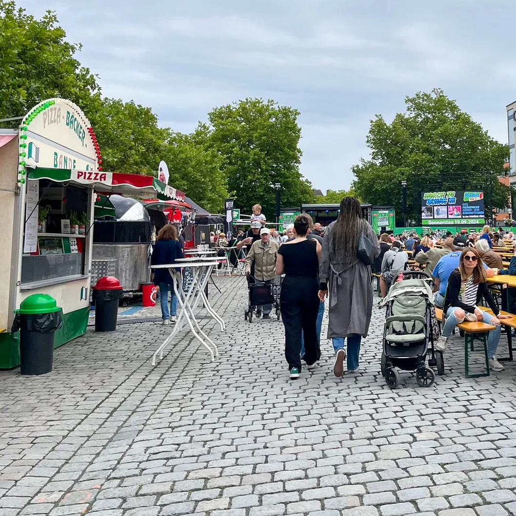 Menschen sitzen an langen Holzbänken und -tischen auf einem gepflasterten Platz neben Essensständen.