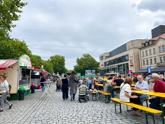 Menschen sitzen an langen Holzbänken und -tischen auf einem gepflasterten Platz neben Essensständen. Klick öffnet Veranstaltungstipp. 