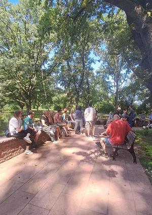 Gruppe von Menschen sitzt und steht an einem sonnigen Tag auf einer gepflasterten Fläche unter großen Bäumen neben einem Gewässer.