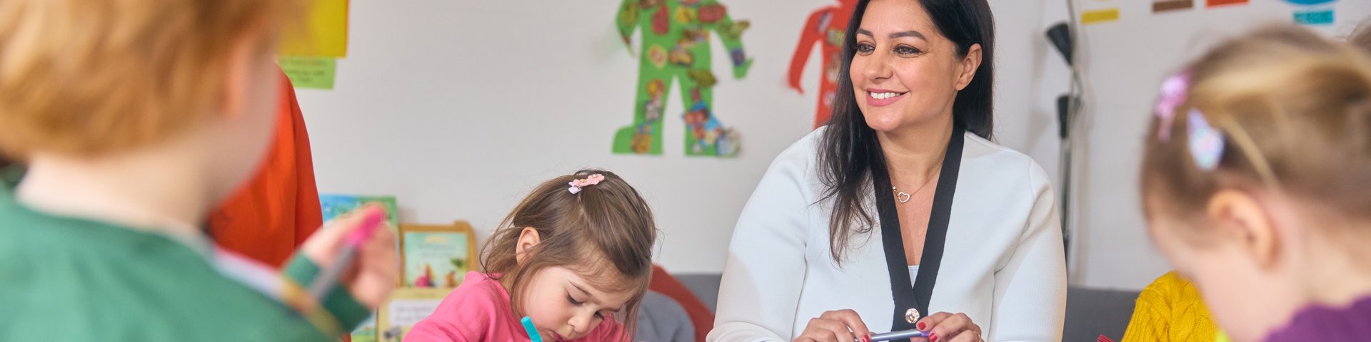 Kinder sitzen an einem Tisch mit Bastelmaterialien und der Erzieherin in einem Raum mit bunten Kinderzeichnungen an der Wand.