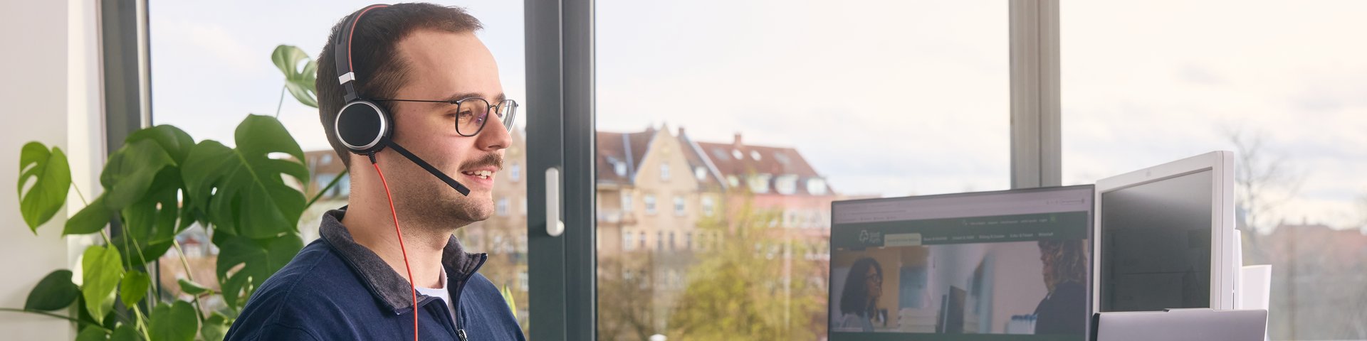 Junger Mann mit Headset steht am Schreibtisch vor zwei Monitoren in einem hellem Büro mit Blick auf Häuser.