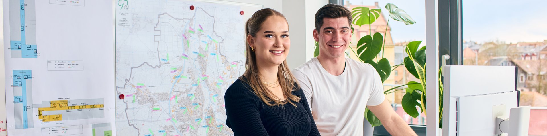 Zwei Personen stehen an einem Schreibtisch mit mehreren Bildschirmen und einem Laptop in einem Büro mit großen Fenstern und einer Wandkarte.