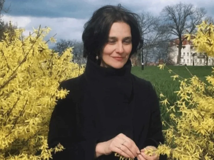 Katja-Petrowskaja mit dunkler Kleidung steht zwischen blühenden gelben Sträuchern auf einer Wiese vor bewölktem Himmel. Klick öffnet Veranstaltungstipp.