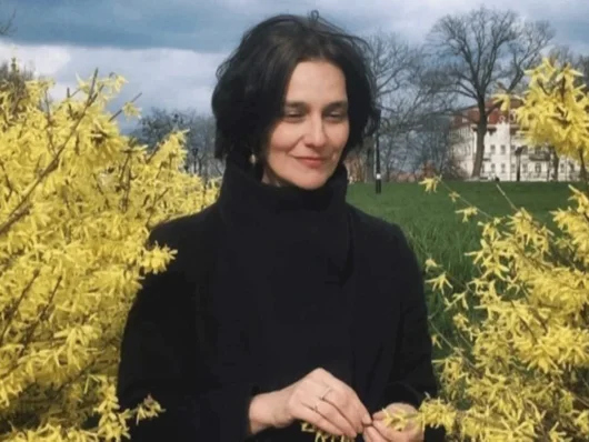 Katja-Petrowskaja mit dunkler Kleidung steht zwischen blühenden gelben Sträuchern auf einer Wiese vor bewölktem Himmel Klick öffnet die Meldung.