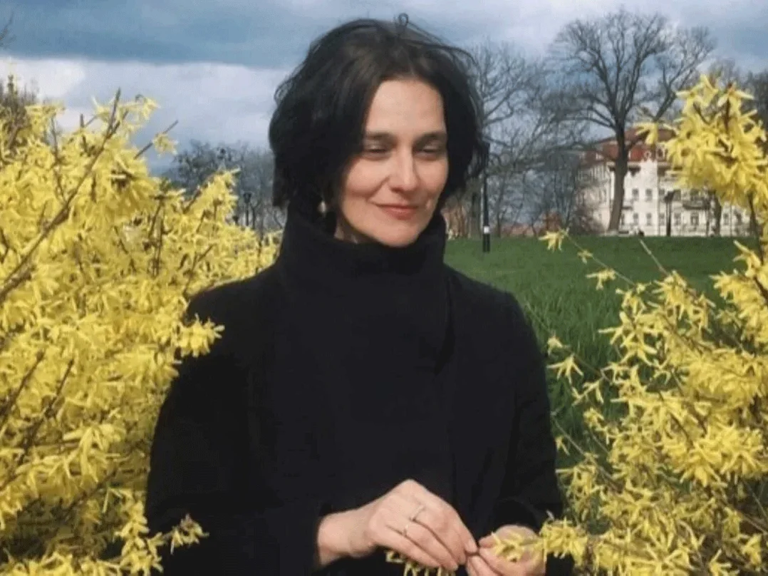 Katja-Petrowskaja mit dunkler Kleidung steht zwischen blühenden gelben Sträuchern auf einer Wiese vor bewölktem Himmel