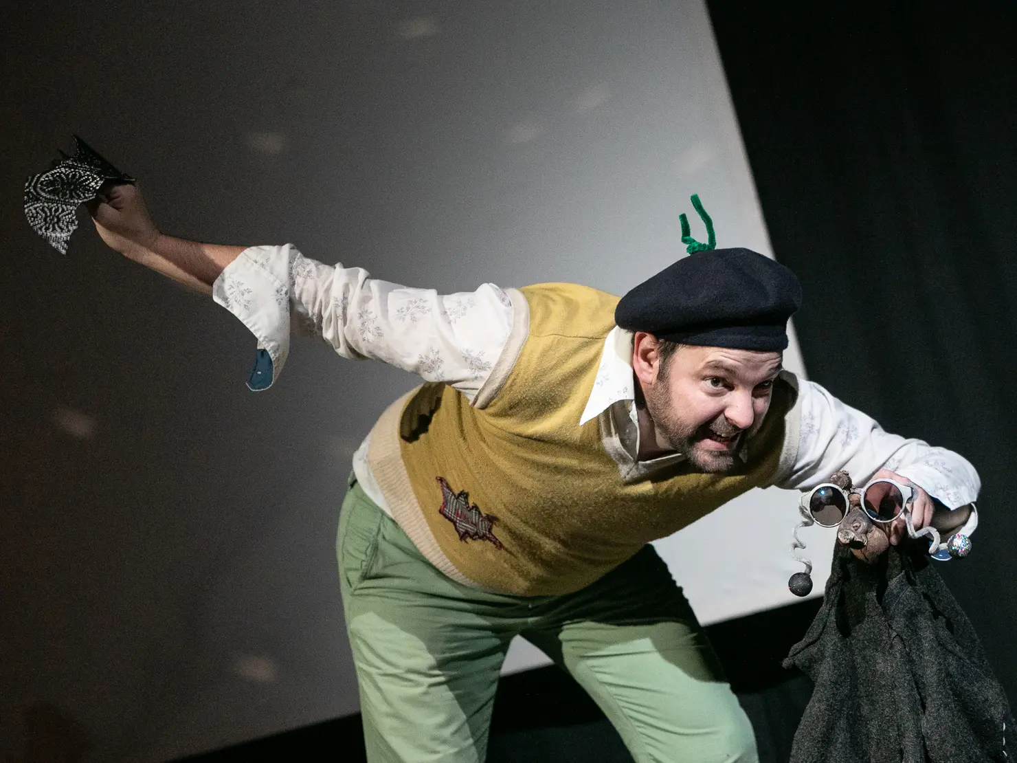 Schauspieler in einem gelben Pullover und grüner Hose, der dynamisch auf einer Bühne posiert. Er trägt eine schwarze Mütze, hält eine schwarze Jacke und eine Sonnebrille. Der Gesichtsausdruck ist lebhaft und ausdrucksvoll.
