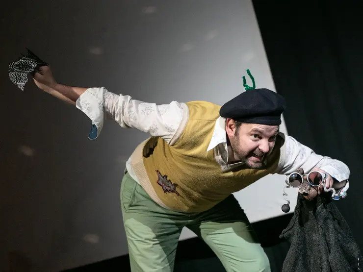 Schauspieler in einem gelben Pullover und grüner Hose, der dynamisch auf einer Bühne posiert. Er trägt eine schwarze Mütze, hält eine schwarze Jacke und eine Sonnebrille. Der Gesichtsausdruck ist lebhaft und ausdrucksvoll.