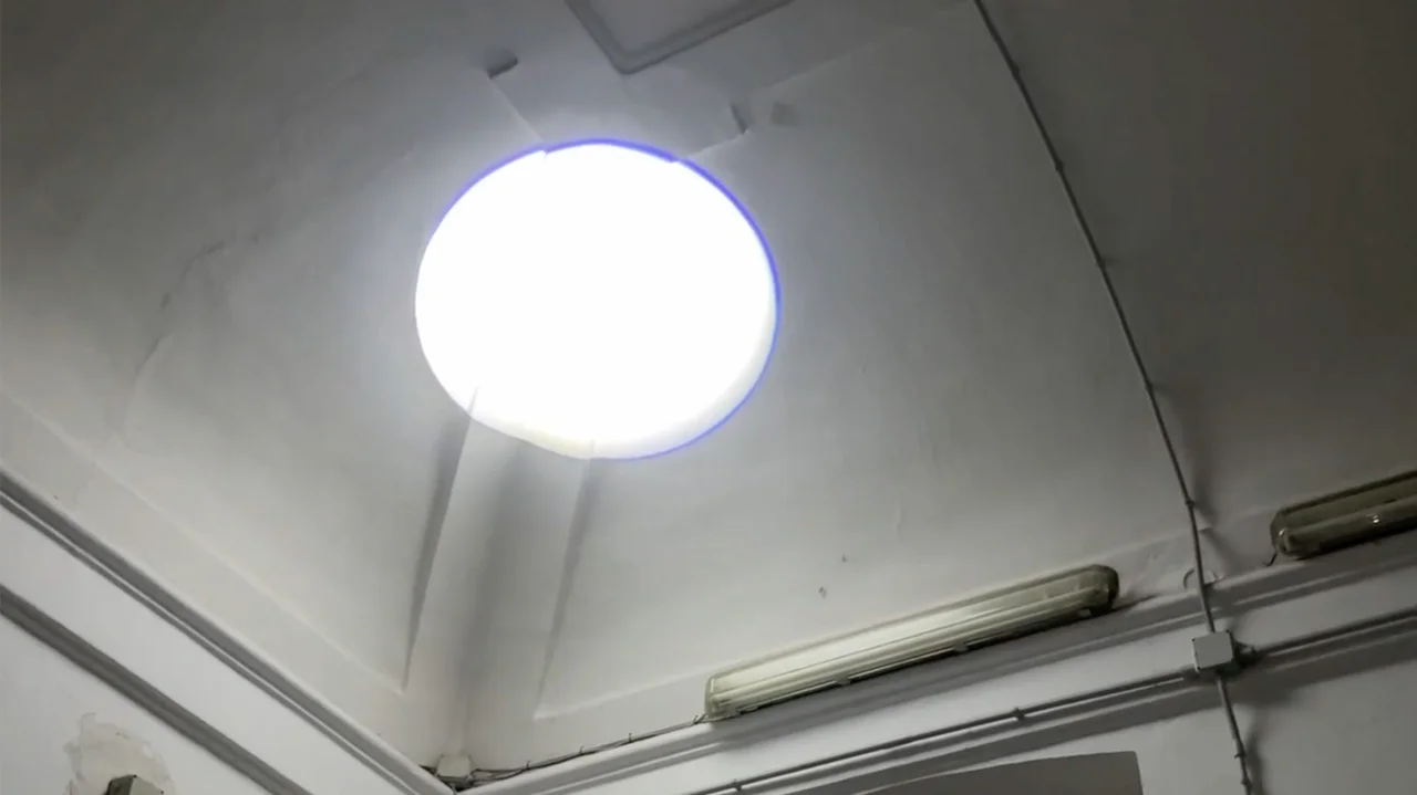 Rundes Oberlicht in einer hohen, weißen Innenraumdecke mit sichtbaren Kabeln und Leuchten an den Wänden