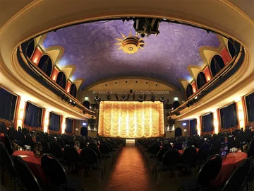 Innenraum der Comödie Fürth mit Bühne, goldener Bühnenvorhang, blauer Decke mit Sonnenornament und Reihen von Tischen mit Stühlen davor.