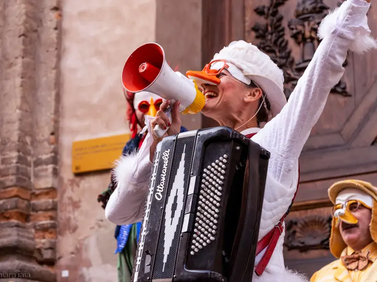 Eine fröhliche Musikerin in einem weißen Kostüm mit Entenmaske spielt Akkordeon und hält ein Megafon in der Hand. Im Hintergrund sind weitere verkleidete Personen zu sehen.