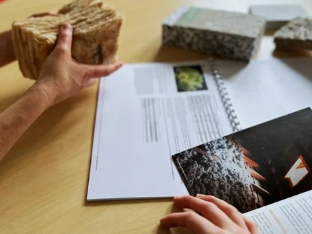 Zwei Hände halten verschiedene Dämmmaterialien über einem Tisch mit geöffneten Broschüren. Klick öffnet die Meldung.