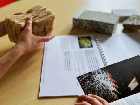 Zwei Hände halten verschiedene Dämmmaterialien über einem Tisch mit geöffneten Broschüren.