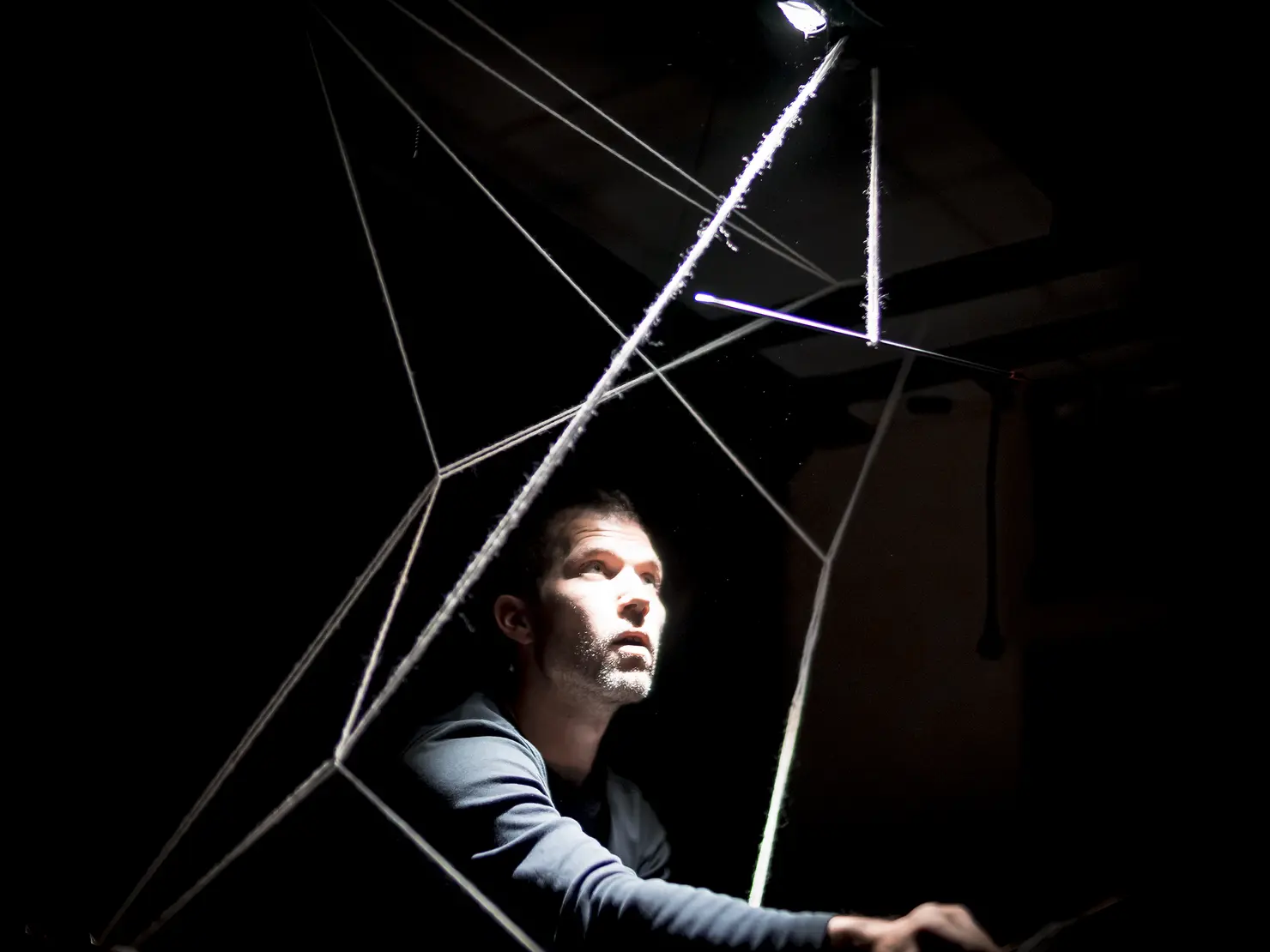 Mann in dunklem Raum, beleuchtet von einem Lichtstrahl, sitzt in einem geometrischen Netz aus Fäden und schaut nach oben.