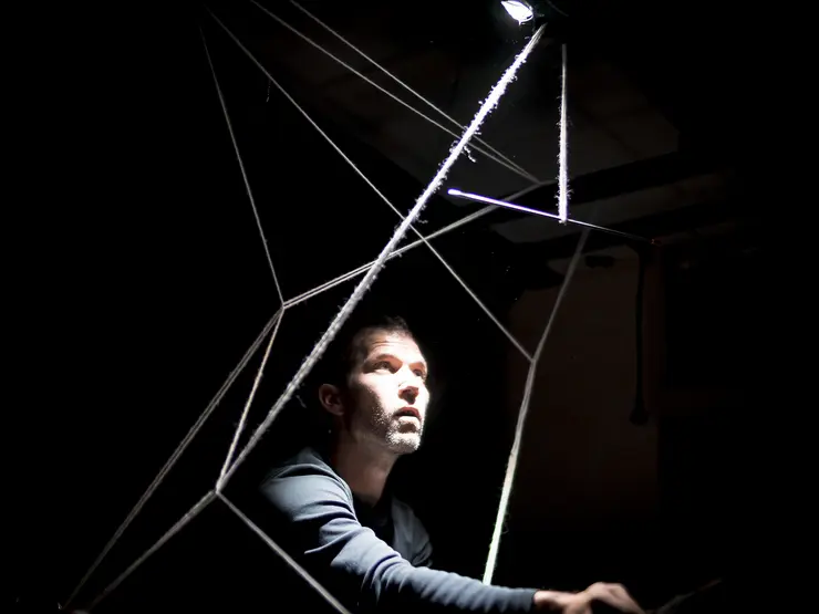 Mann in dunklem Raum, beleuchtet von einem Lichtstrahl, sitzt in einem geometrischen Netz aus Fäden und schaut nach oben.