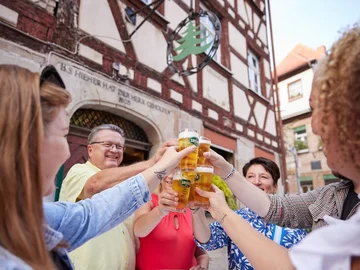 Mehrere Personen stoßen mit Biergläsern vor einem Fachwerkhaus an, das eine Gaststätte ist.