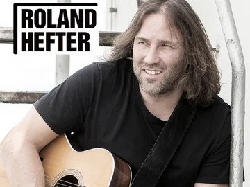 Roland Hefter mit schulterlangem Haar und schwarzem T-Shirt spielt akustische Gitarre vor einer weißen Wand mit dem Schriftzug 'ROLAND HEFTER'