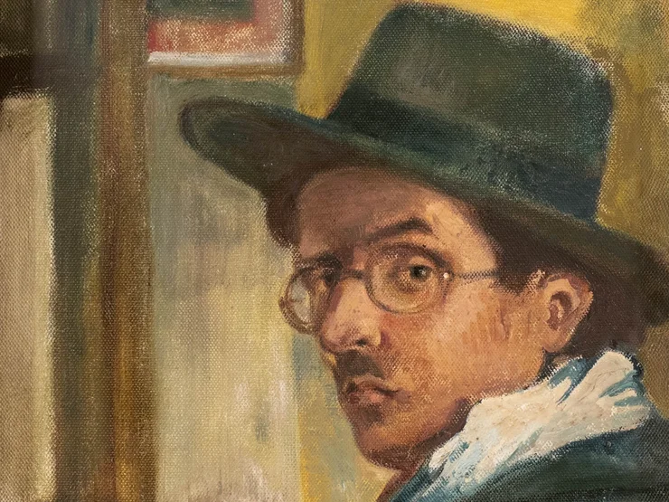 Porträt eines sitzenden Mannes mit Hut und Brille in einem Innenraum