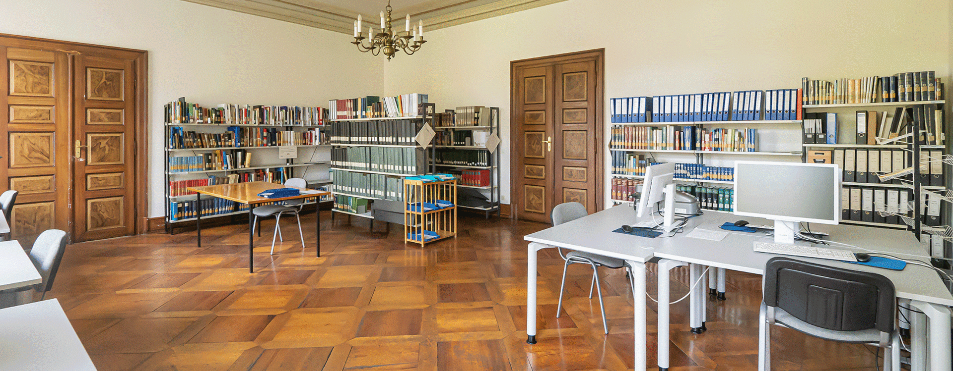 Großer Lesesaal mit Parkettboden, hohen Decken, mehreren Tischen mit Stühlen und Regalen voller Ordner und Bücher an den Wänden.
