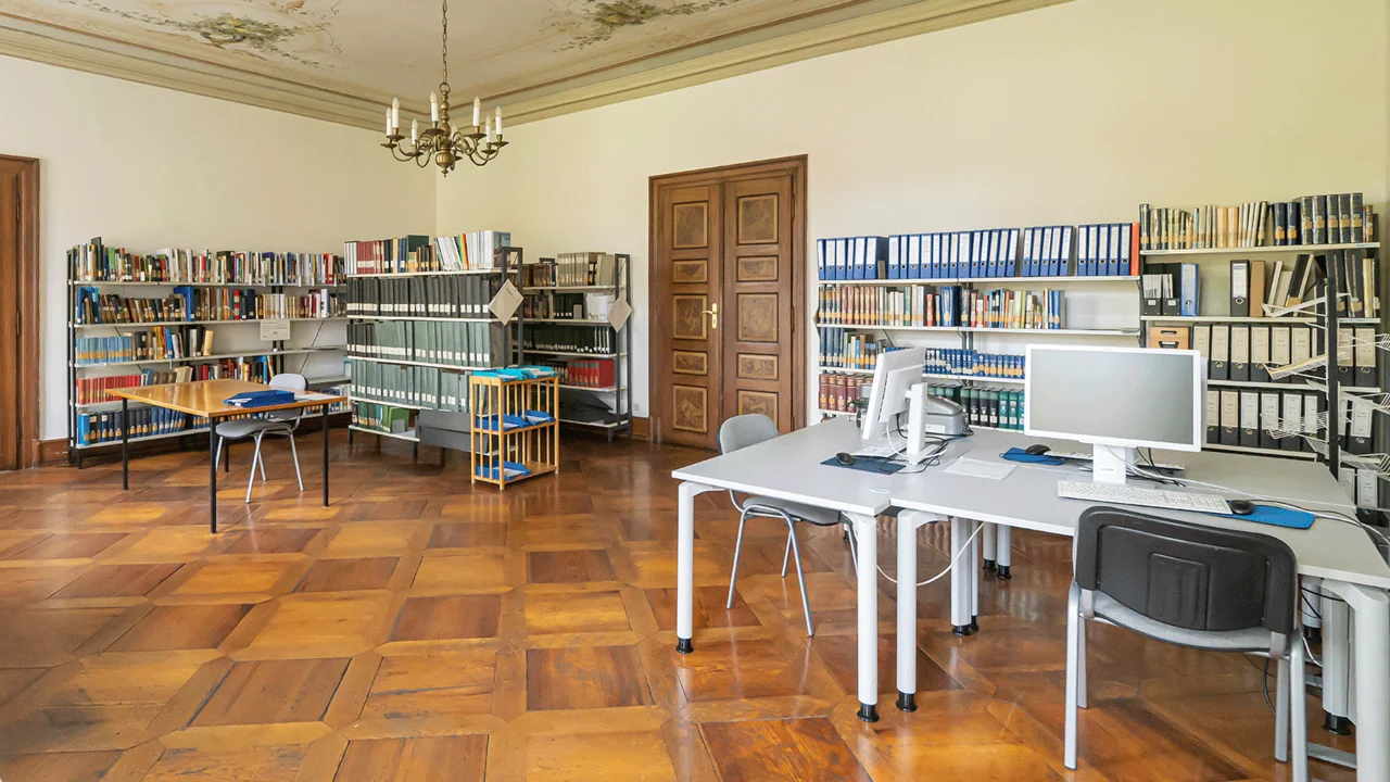 Großer Lesesaal mit Parkettboden, hohen Decken, mehreren Tischen mit Stühlen und Regalen voller Ordner und Bücher an den Wänden.