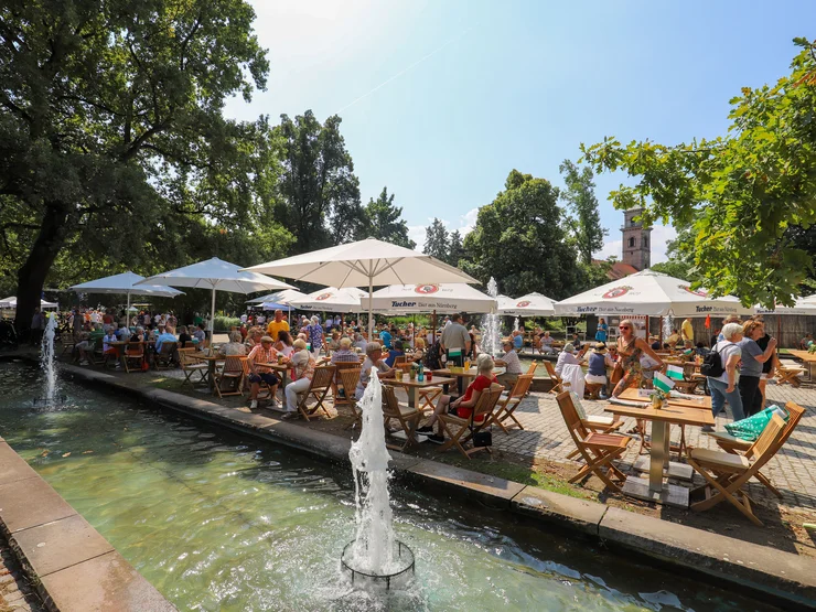 Mehrere Personen sitzen an Holztischen mit Sonnenschirmen neben einem Brunnen im Stadtpark, Klick öffnet Veranstaltungstipp