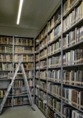 Hohe Regale voller Bücher in einem Archivraum mit einer aufgestellten Metallleiter in der Mitte.