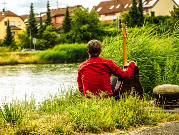 Mann mit Gitarre sitzt an Flussufer.