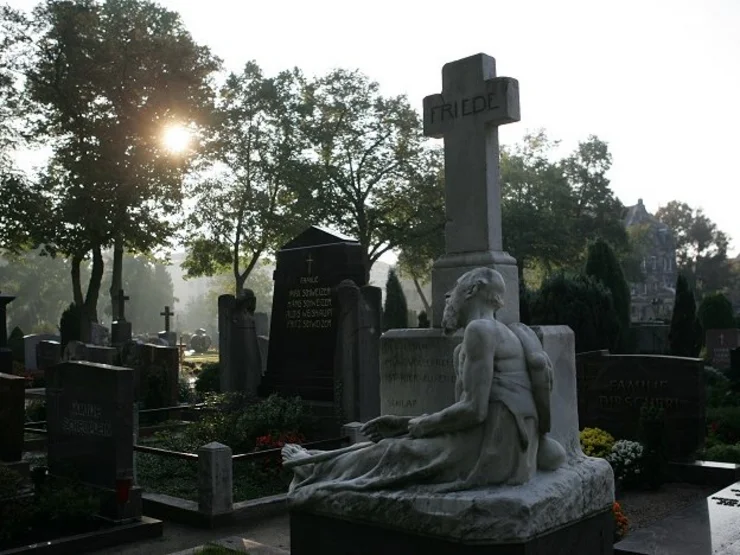 Grabstein mit einer liegenden Skulptur vor einem Kreuz mit der Inschrift 'FRIEDE' auf einem städtischen Friedhof bei Sonnenlicht und Bäumen im Hintergrund.