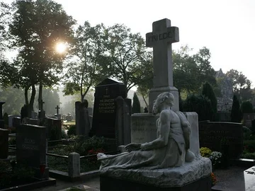 Grabstein mit einer liegenden Skulptur vor einem Kreuz mit der Inschrift 'FRIEDE' auf einem städtischen Friedhof bei Sonnenlicht und Bäumen im Hintergrund.