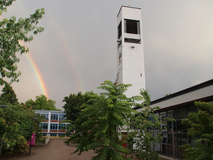 Moderner weißer Kirchturm der Heilig Geist Kirche auf der Hardhöhe dahinter ein Regenbogen am bewölkten Abendhimmel über einem Gemeindezentrum.