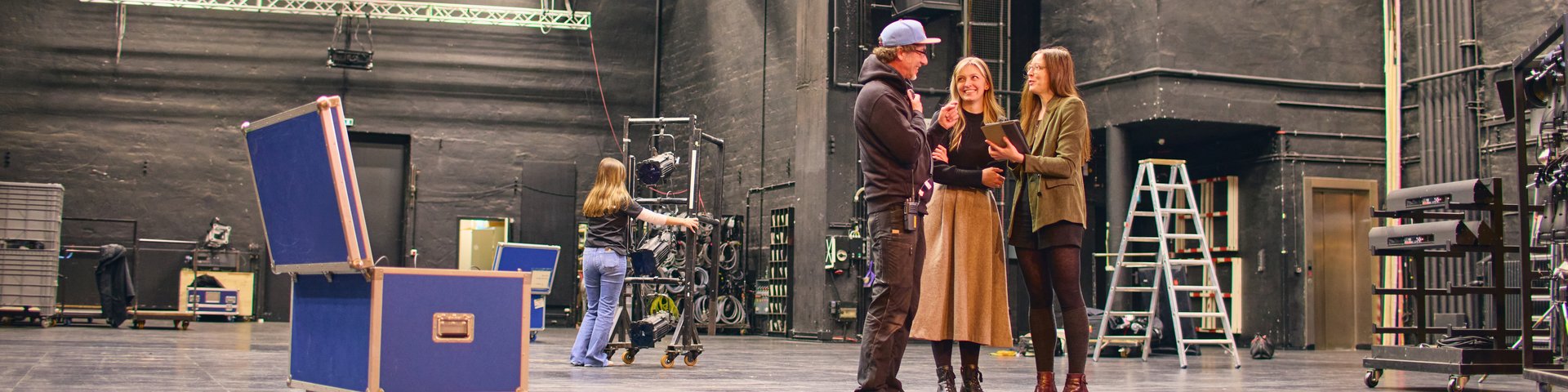 Drei Personen stehen auf einer Bühne in einem Theater, im Hintergrund sind technische Bühnenelemente zu sehen.