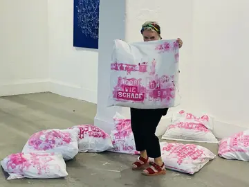 Foto in einer Ausstellung. Die Künstlerin hält einen mit pinker Schrift bedruckten Kissenbezug hoch. Er ist Teil der Installation "Letzte Worte", die hinter ihr in der Ausstellung aufgebaut ist