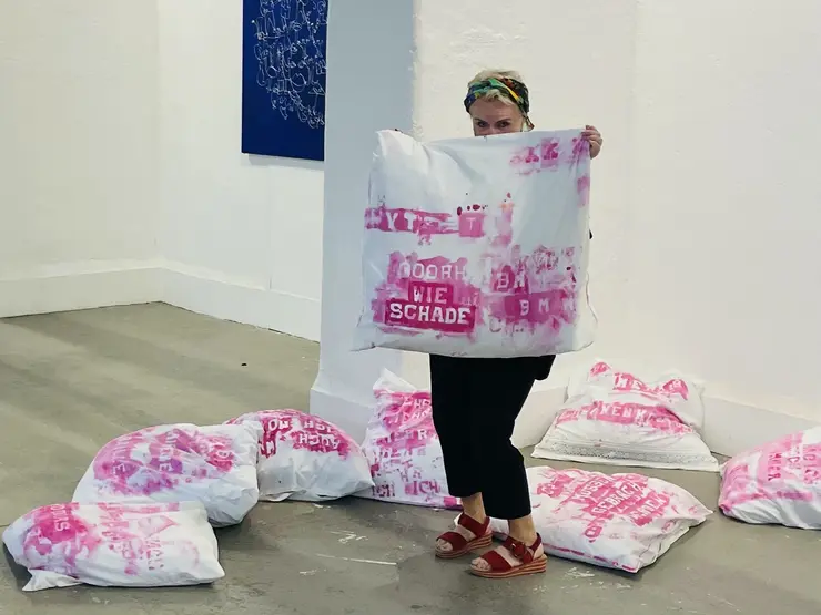 Foto in einer Ausstellung. Die Künstlerin hält einen mit pinker Schrift bedruckten Kissenbezug hoch. Er ist Teil der Installation "Letzte Worte", die hinter ihr in der Ausstellung aufgebaut ist