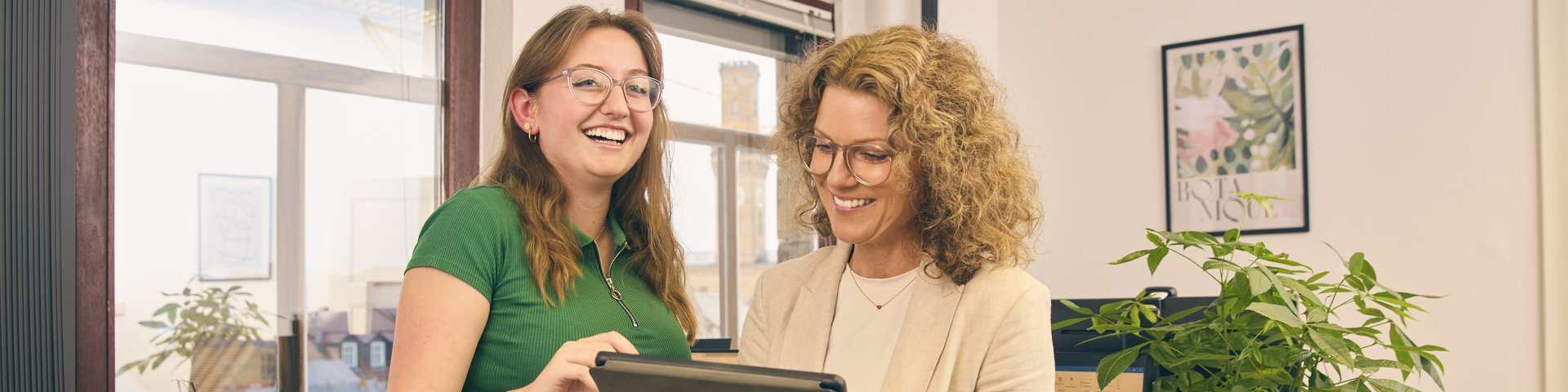 Zwei Frauen stehen in einem Büro vor einem Schreibtisch und blicken auf ein Tablet.