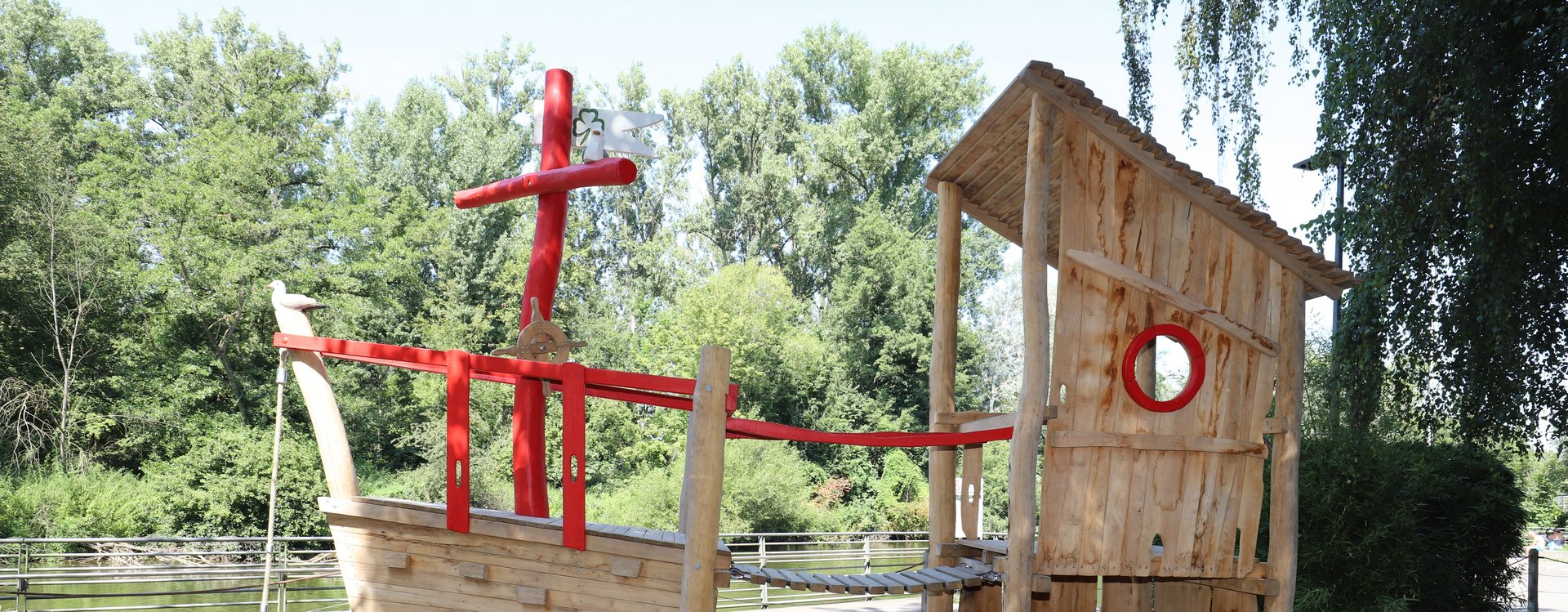 Spielplatz Uferpromenade Piratenschiff aus Holz vor der Rednitz und Bäumen.