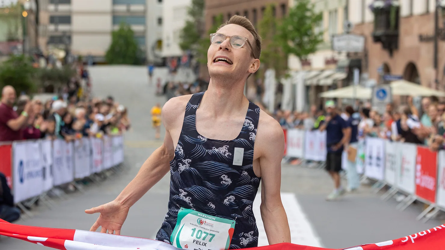 Mann in Sportbekleidung überquert als Erster die Ziellinie und durchläuft ein rot-weißes Zielband.