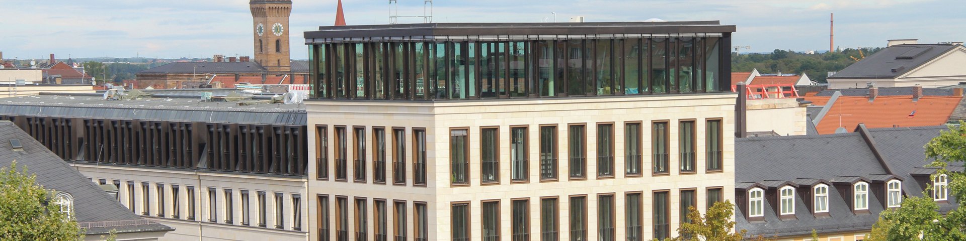 Blick auf ein modernes mehrstöckiges Gebäude mit großen Fenstern in einer belebten Innenstadtumgebung mit Bäumen und Menschen auf dem Platz.