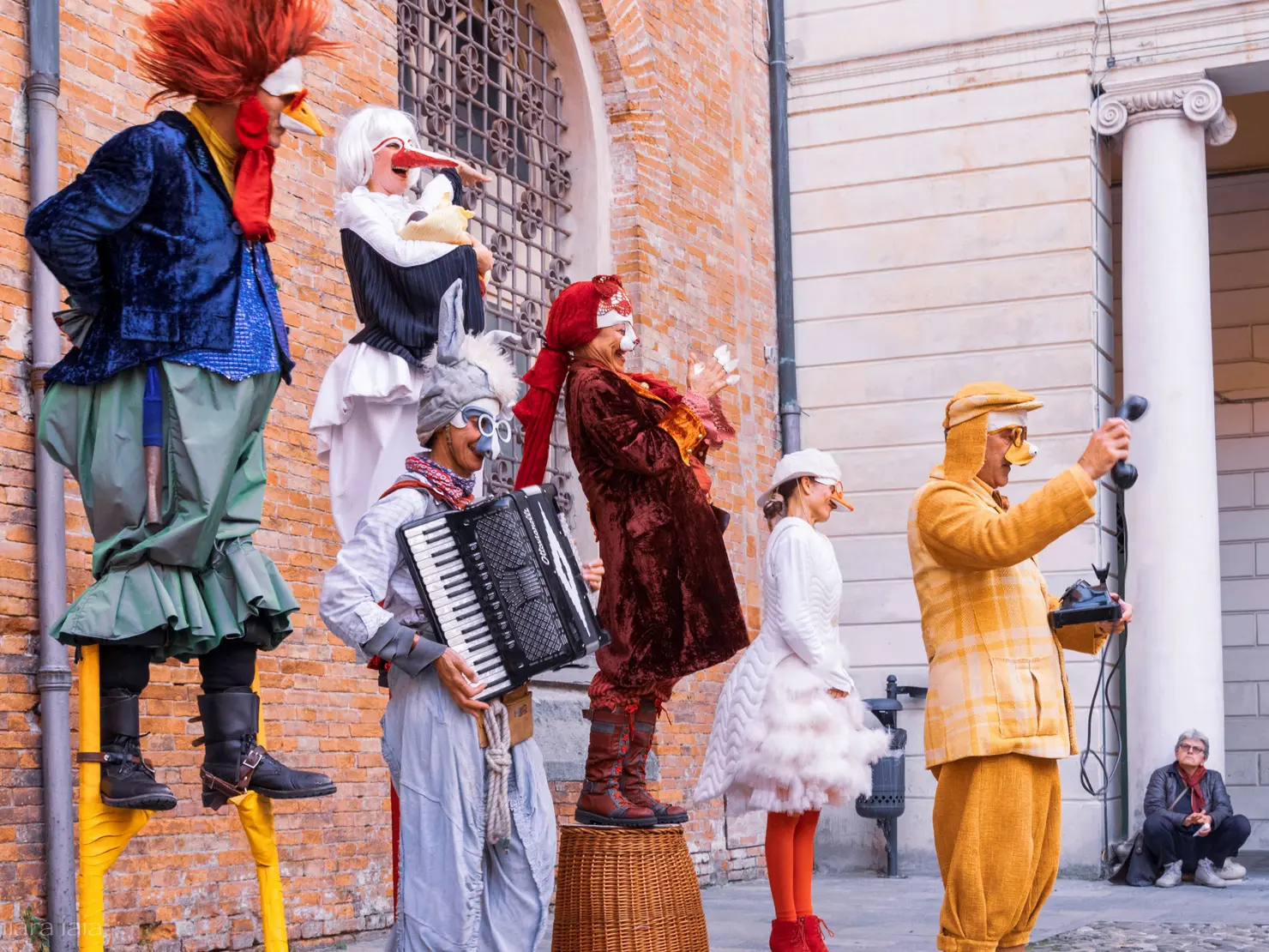 Sechs Künstler*innen in bunten Kostümen und Masken führen eine Straßenshow auf. Zwei stehen auf hohen Stelzen, während andere Instrumente spielen und singen.