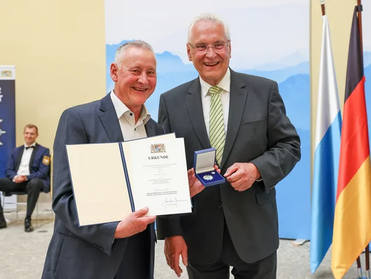 OB Jung hält Urkunde und Stern der Sicherheit als Anstecknadel in einem kleinen Box in der Hand. Neben ihm Innenminister Herrmann. Klick öffnet die Meldung.