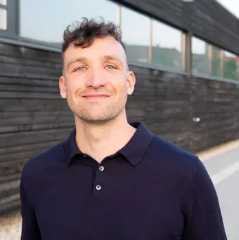 Mann in blauem Hemd vor Holzhaus. Klick öffnet Bild in größerer Ansicht.