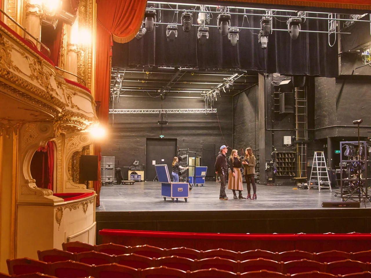 Bühnenraum eines Theaters mit rotem Vorhang, goldverzierten Logen, roten Sitzreihen und drei Personen auf der Bühne mit technischer Ausstattung.