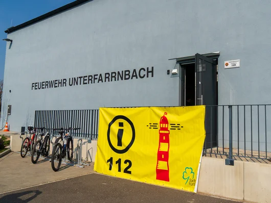 Gebäude der Feuerwehr Unterfarrnbach, behängt mit einem gelben Banner, auf dem ein roter Leuchtturm, die Notrufnummer 112 und das Logo der Stadt Fürth zu sehen ist. Klick öffnet die Meldung.