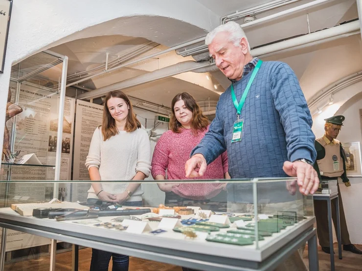Drei Personen stehen vor einer Vitrine mit verschiedenen Exponaten im Kriminalmuseum, eine Person zeigt auf die Ausstellungsstücke.