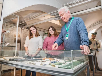 Drei Personen stehen vor einer Vitrine mit verschiedenen Exponaten im Kriminalmuseum, eine Person zeigt auf die Ausstellungsstücke.