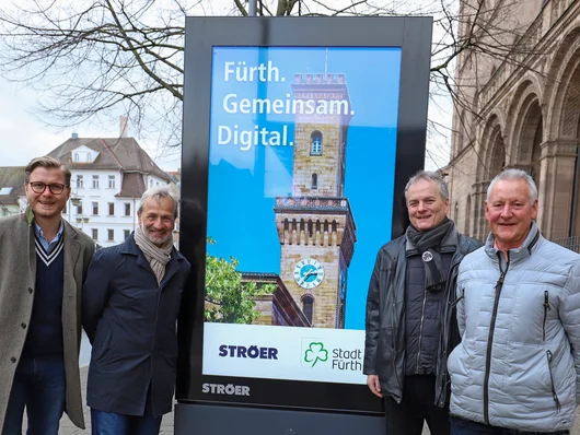 Die Mitwirkenden an den Cityscreens stehen in der Nähe des Rathauses zusammen mit dem Wirtschaftsreferent und dem Oberbürgermeister vor einem großen Bildschirm. Klick öffnet die Meldung.
